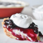 A close-up of a slice of blueberry cream cheese pie with a graham cracker crust. The pie is topped with a glossy blueberry sauce and a dollop of whipped cream.