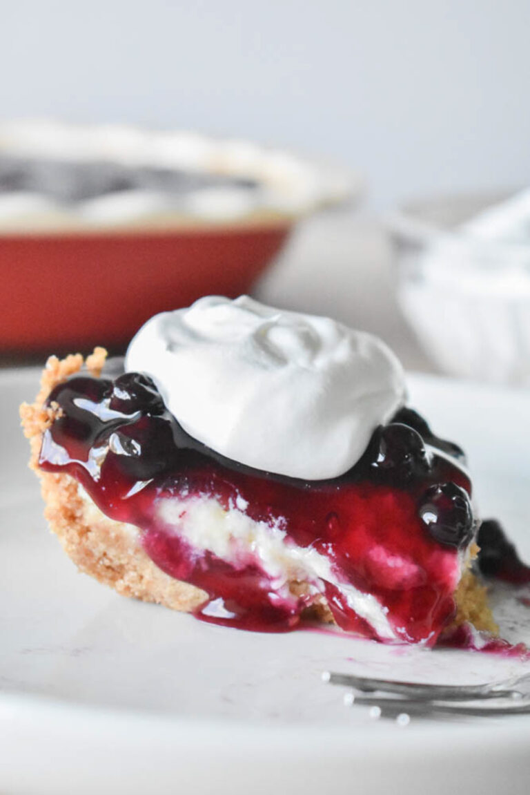 A close-up of a slice of blueberry cream cheese pie with a graham cracker crust. The pie is topped with a glossy blueberry sauce and a dollop of whipped cream.