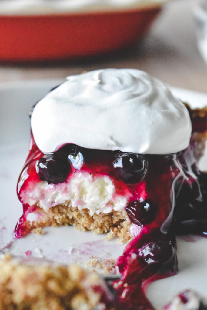 A slice of blueberry cream cheese pie with a crumbly base, topped with glossy blueberry sauce and a swirl of whipped cream.