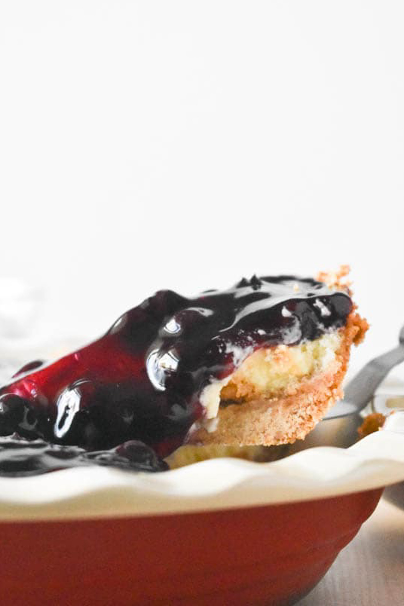 A slice of blueberry cream cheese pie being lifted out of a red pie pan. The pie has a crumbly crust topped with glossy, dark purple blueberry glaze.