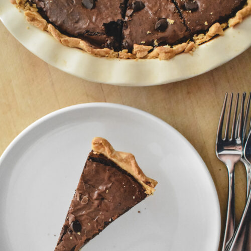 A slice of rich brownie pie with a flaky crust on a white plate next to two forks. Above, the remaining pie is in a white dish, garnished with chocolate chips.