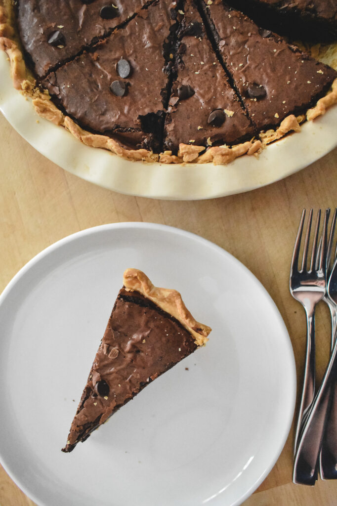 A slice of rich brownie pie with a flaky crust on a white plate next to two forks. Above, the remaining pie is in a white dish, garnished with chocolate chips.
