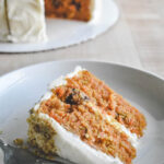 A slice of carrot cake with cream cheese frosting on a white plate, accompanied by a fork. The cake is moist and layered with visible carrots and nuts, with a whole cake in the background.