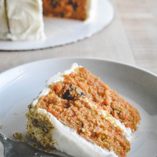A slice of carrot cake with cream cheese frosting on a white plate, accompanied by a fork. The cake is moist and layered with visible carrots and nuts, with a whole cake in the background.