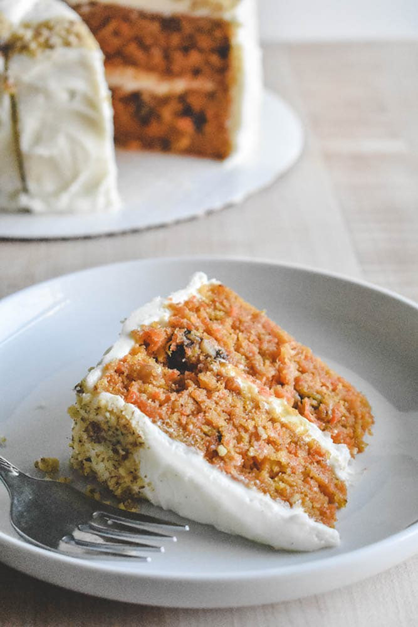 A slice of carrot cake with cream cheese frosting on a white plate, accompanied by a fork. The cake is moist and layered with visible carrots and nuts, with a whole cake in the background.