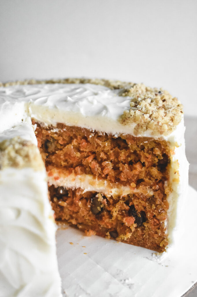 Side view of a round carrot cake with a slice missing. Moist layers with nuts and cream cheese frosting can be seen. The cake is topped with chopped nuts along the edge.