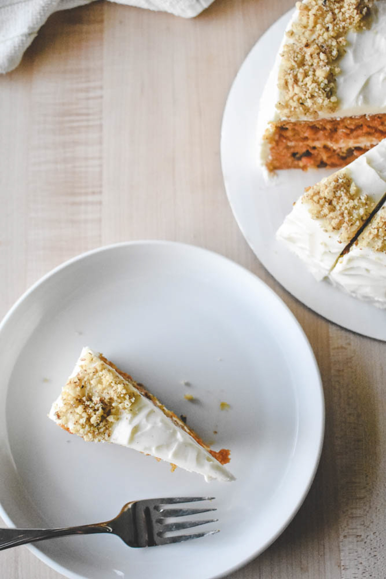 A slice of carrot cake with white frosting and chopped nuts on a white plate with a fork. The rest of the cake is partly visible on a wooden table.
