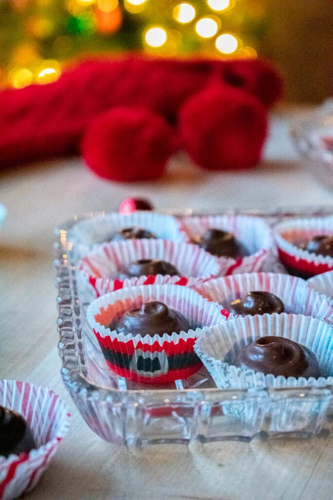 Festive cherry cordials in red Christmas paper candy cups sit in a glass dish on a wooden table. Warm holiday lights and red decor are in the background