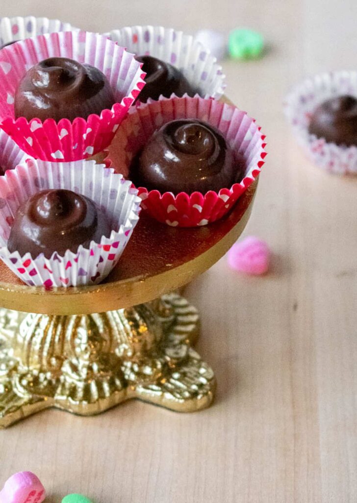 Cherry cordials in red and white paper cups sit on a decorative golden stand. Colorful candy hearts are scattered on a wooden surface.