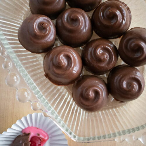 Ten smooth, swirl-topped cherry cordials on a glass plate. One is bitten, revealing a bright pink filling with a cherry, in a paper cup.