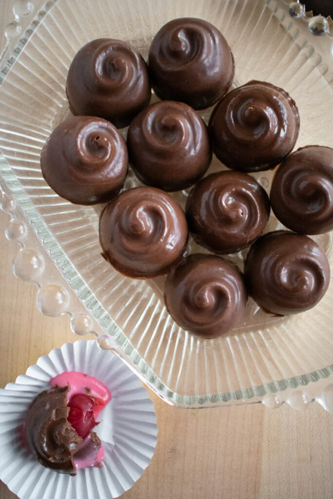 Ten smooth, swirl-topped cherry cordials on a glass plate. One is bitten, revealing a bright pink filling with a cherry, in a paper cup.