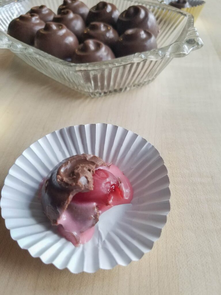 A bitten chocolate covered cherry with a pink cherry filling sits in a paper cup on a wooden surface. In the background, a dish holds more chocolate candies.