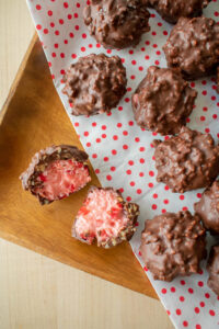 Close up of homemade cherry mash candies with nutty, textured exteriors on polka-dotted paper. One candy is cut open, revealing a pink, creamy filling.