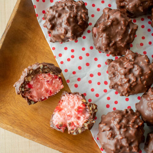 Close up of homemade cherry mash candies with nutty, textured exteriors on polka-dotted paper. One candy is cut open, revealing a pink, creamy filling.