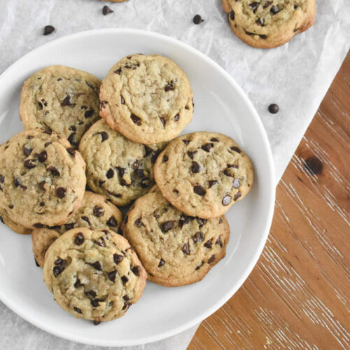 A white plate filled with golden brown chewy chocolate chip cookies sits on parchment paper. Additional cookies are scattered on a rustic wooden table.
