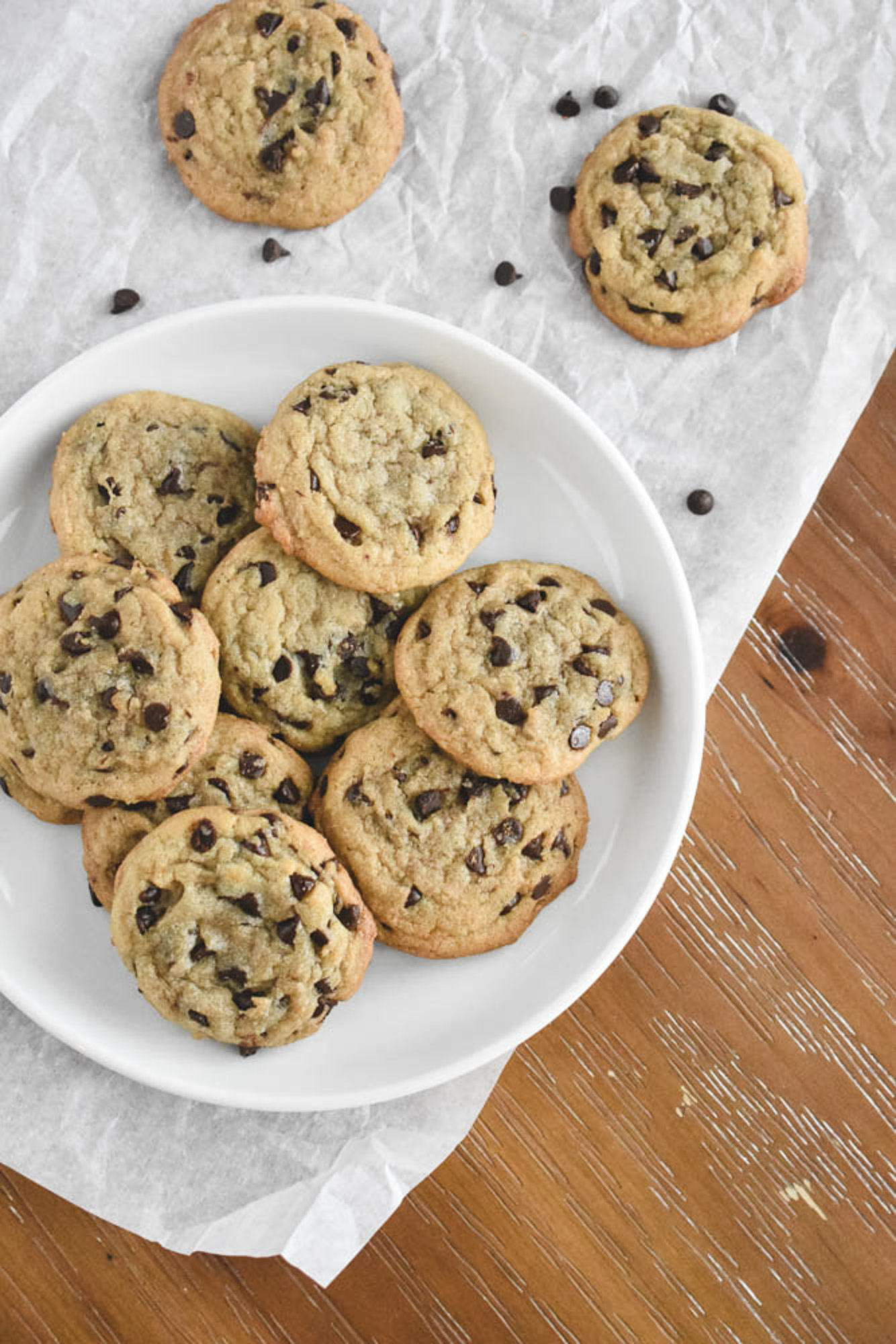 A white plate filled with golden brown chewy chocolate chip cookies sits on parchment paper. Additional cookies are scattered on a rustic wooden table.