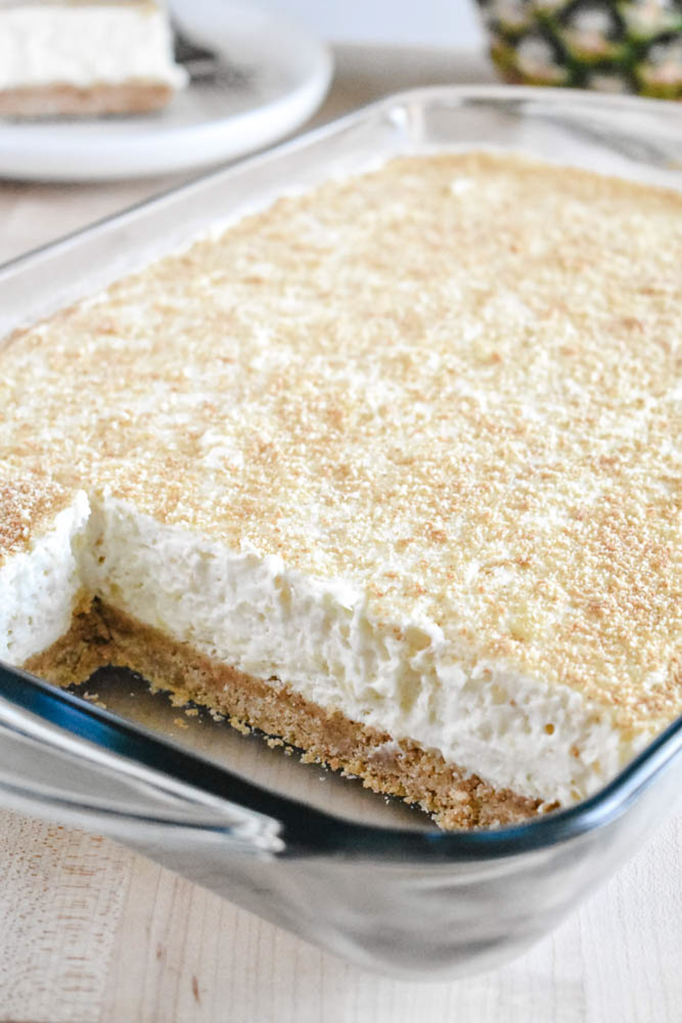 A rectangular glass dish with creamy, thick pineapple dessert on top of a light golden crust. A slice is removed, showing the textured layers inside.