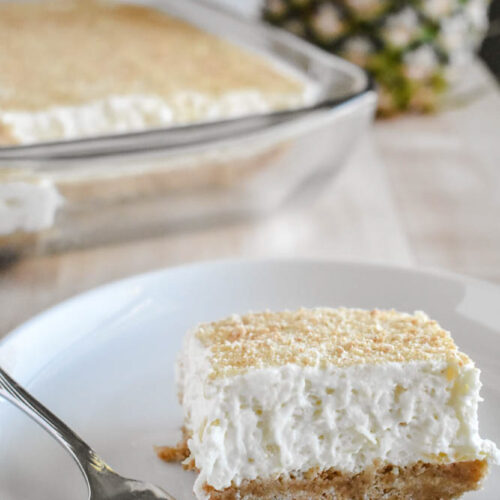 A square slice of creamy pineapple dessert with a graham cracker crust sits on a white plate next to a fork. In the background, a whole pineapple and a glass baking dish with more dessert are visible.