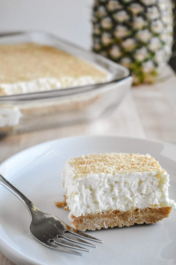A square slice of creamy pineapple dessert with a graham cracker crust sits on a white plate next to a fork. In the background, a whole pineapple and a glass baking dish with more dessert are visible.