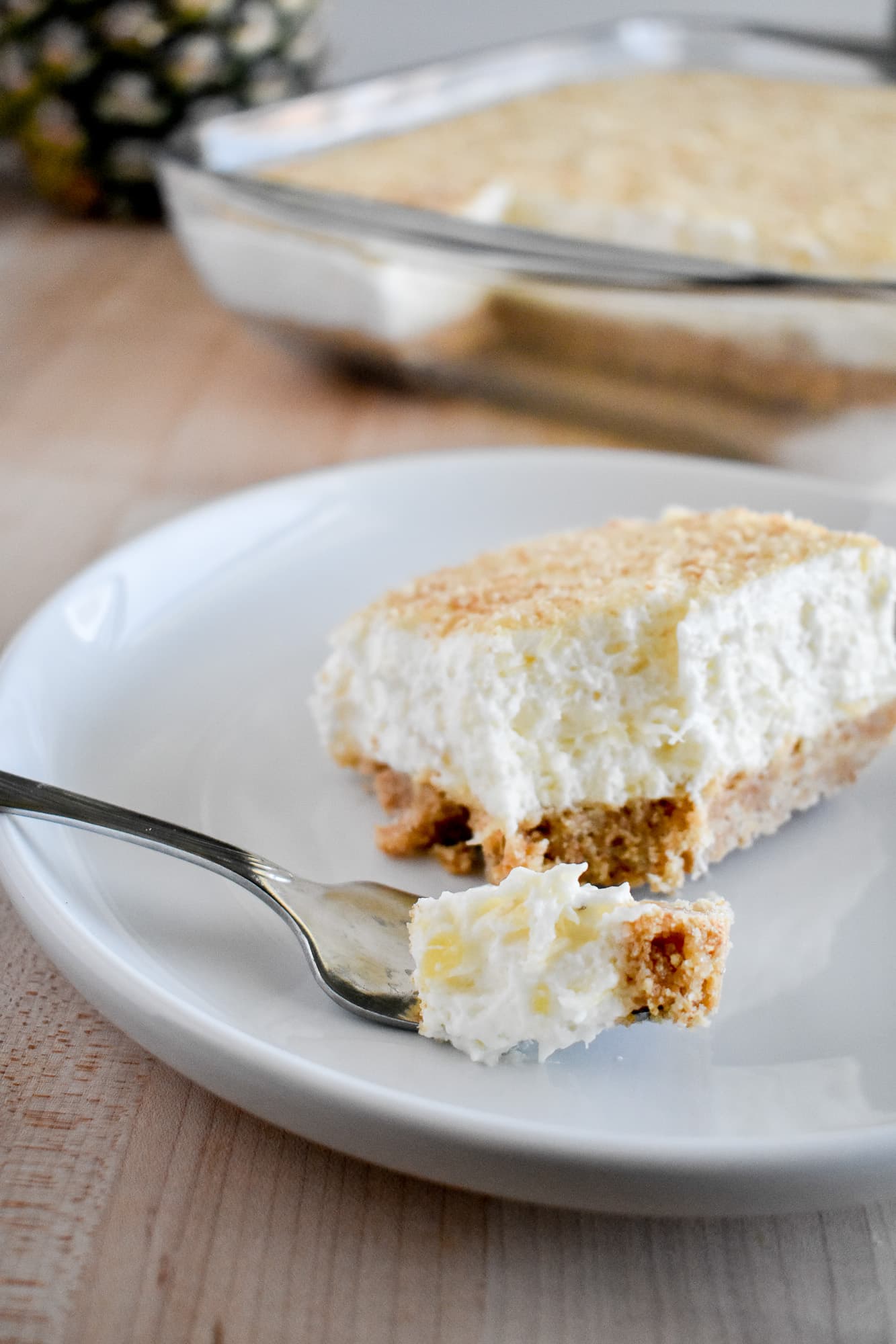 A piece of creamy pineapple dessert sits on a white plate, featuring a crumbly crust and light whipped topping. A fork holds a small bite. A larger dish is in the background.