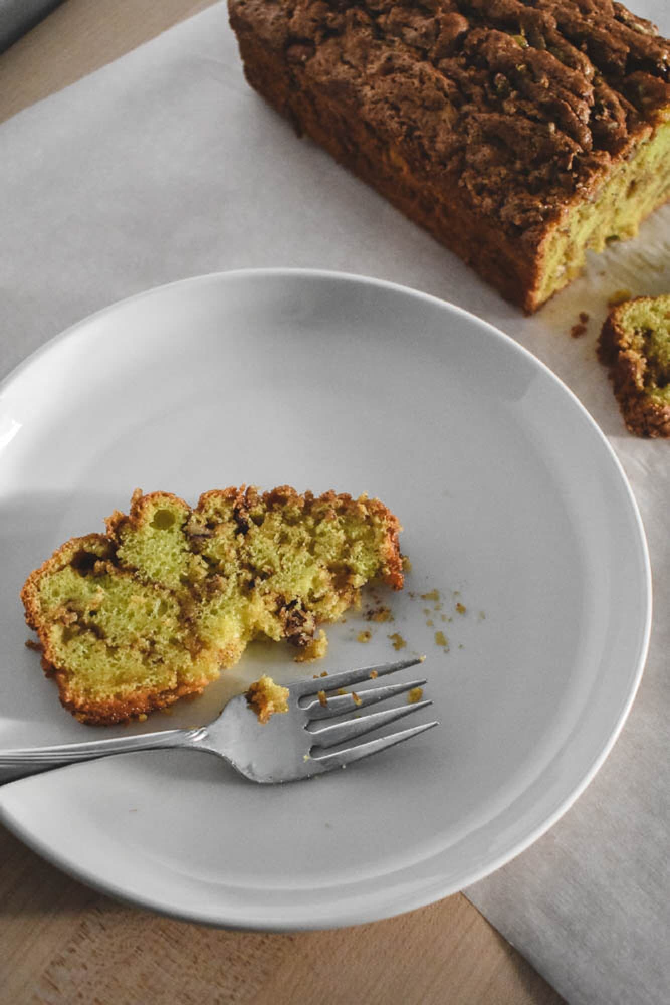 A slice of pistachio bread with pecan bits visible on a white plate and fork. Nearby, a loaf reveals a golden brown exterior. 
