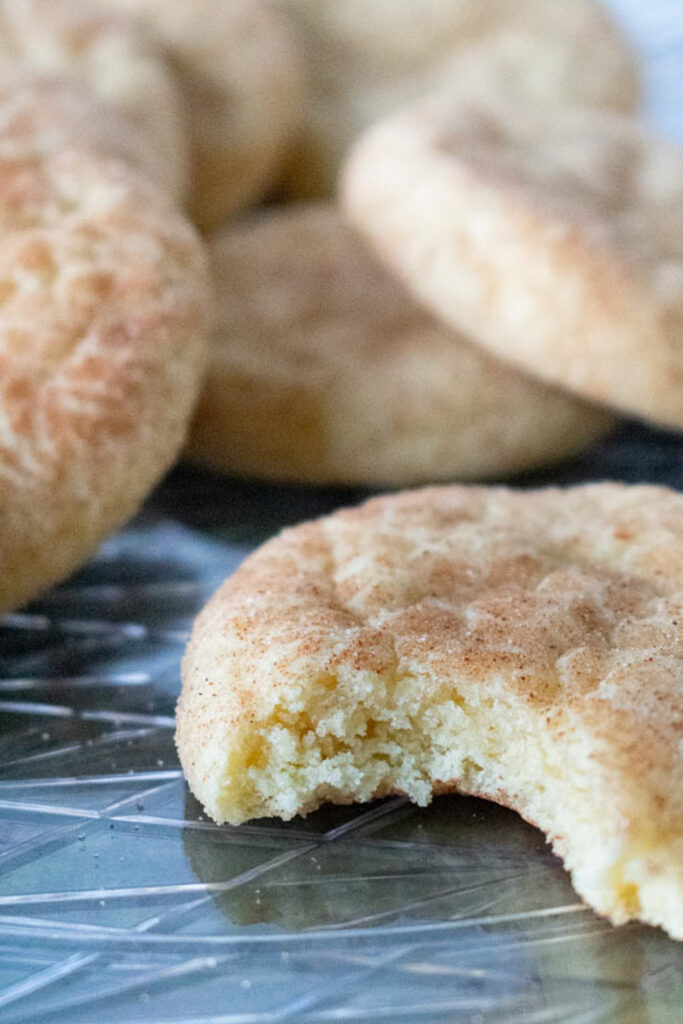 Close up of a snickerdoodle cookie with a bite taken out, showing a soft interior. Other cookies are blurred in the background, on a patterned surface.