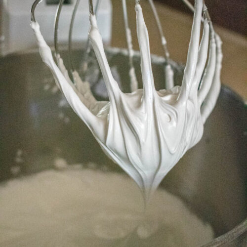 A metal whisk attachment, covered in fluffy white royal icing, is held above a mixing bowl.
