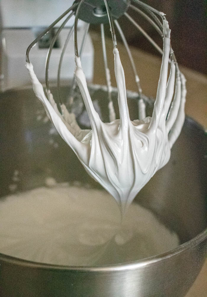 A metal whisk attachment, covered in fluffy white royal icing, is held above a mixing bowl.