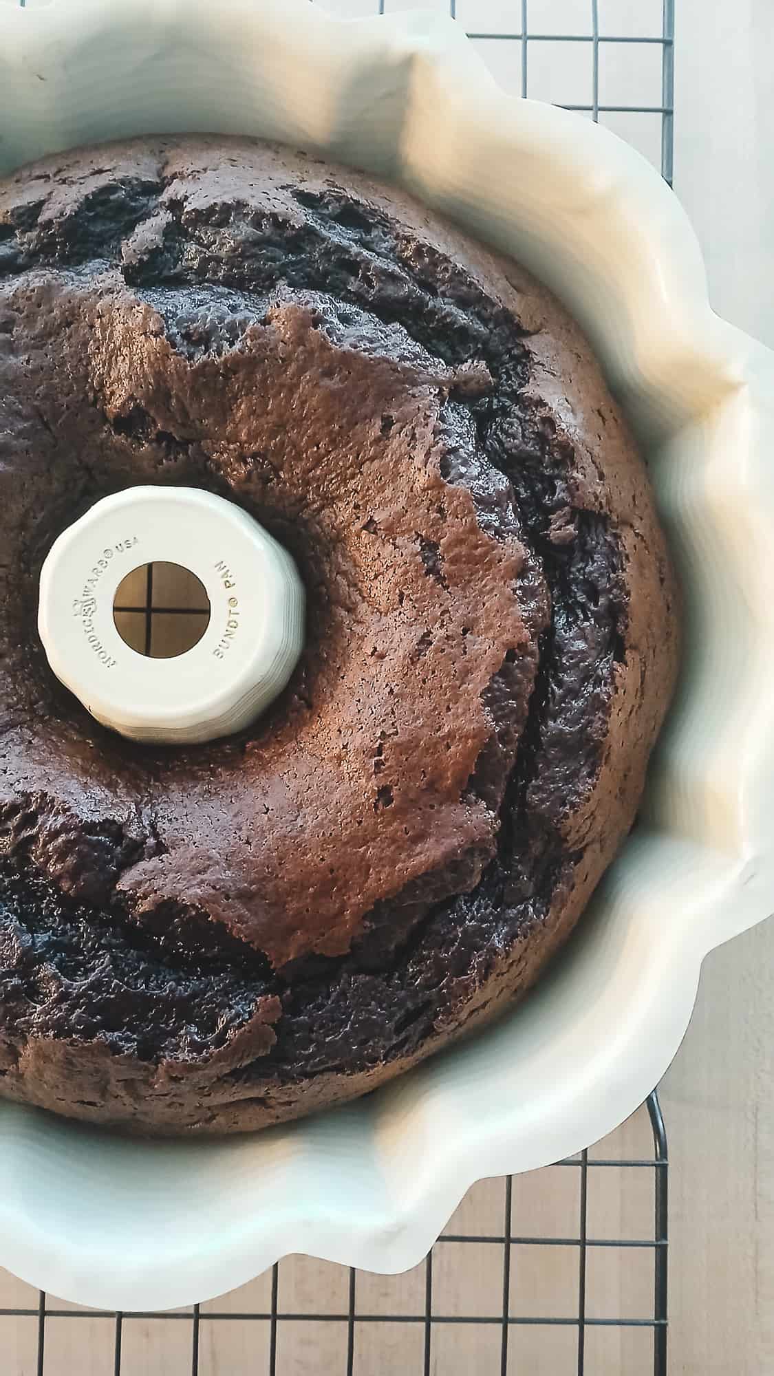 A freshly baked chocolate Black Russian Bundt cake cooling in a white pan on a wire rack. The rich, dark crust and slightly cracked surface show the moist, homemade texture of the cake before it is inverted