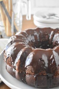 A chocolate Bundt cake (Black Russian Cake) with a glossy chocolate glaze sits on a white plate. The background is blurred, showcasing dishes and utensils.