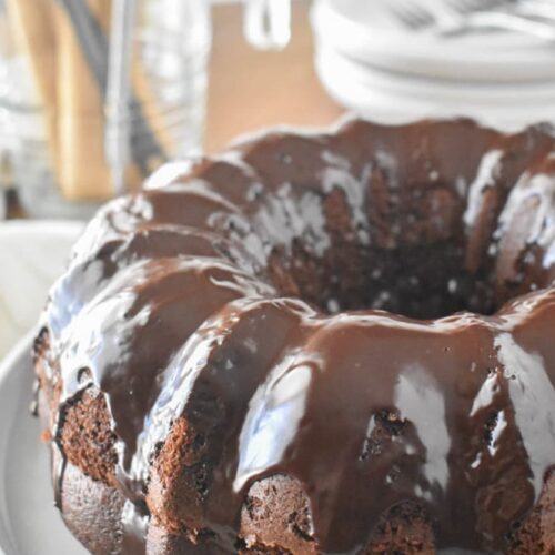 A chocolate Bundt cake (Black Russian Cake) with a glossy chocolate glaze sits on a white plate. The background is blurred, showcasing dishes and utensils.