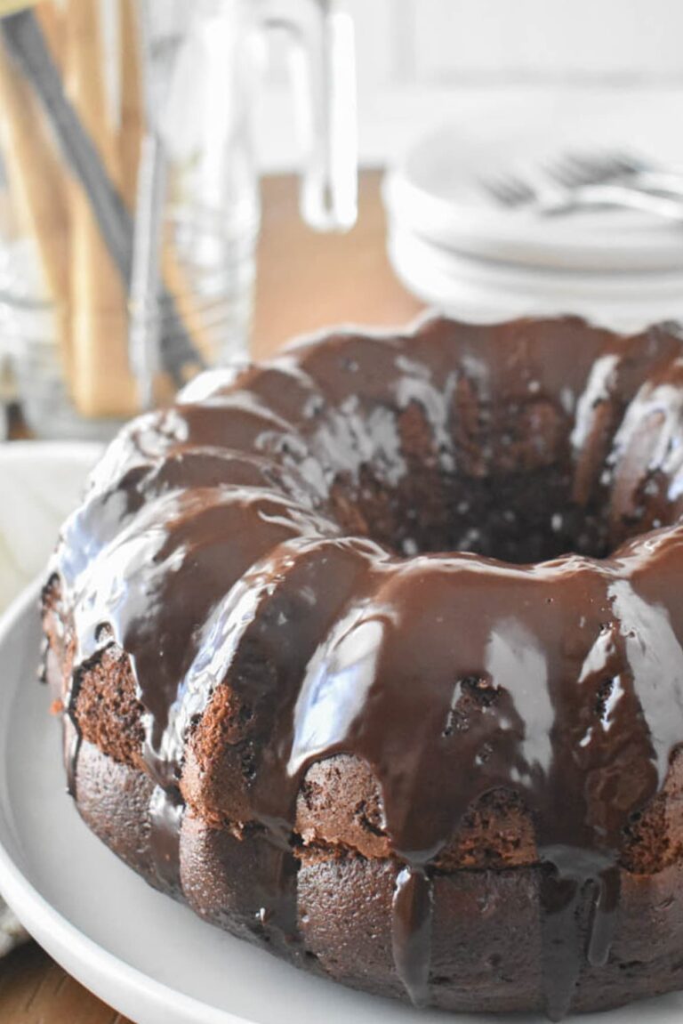 A chocolate Bundt cake (Black Russian Cake) with a glossy chocolate glaze sits on a white plate. The background is blurred, showcasing dishes and utensils.