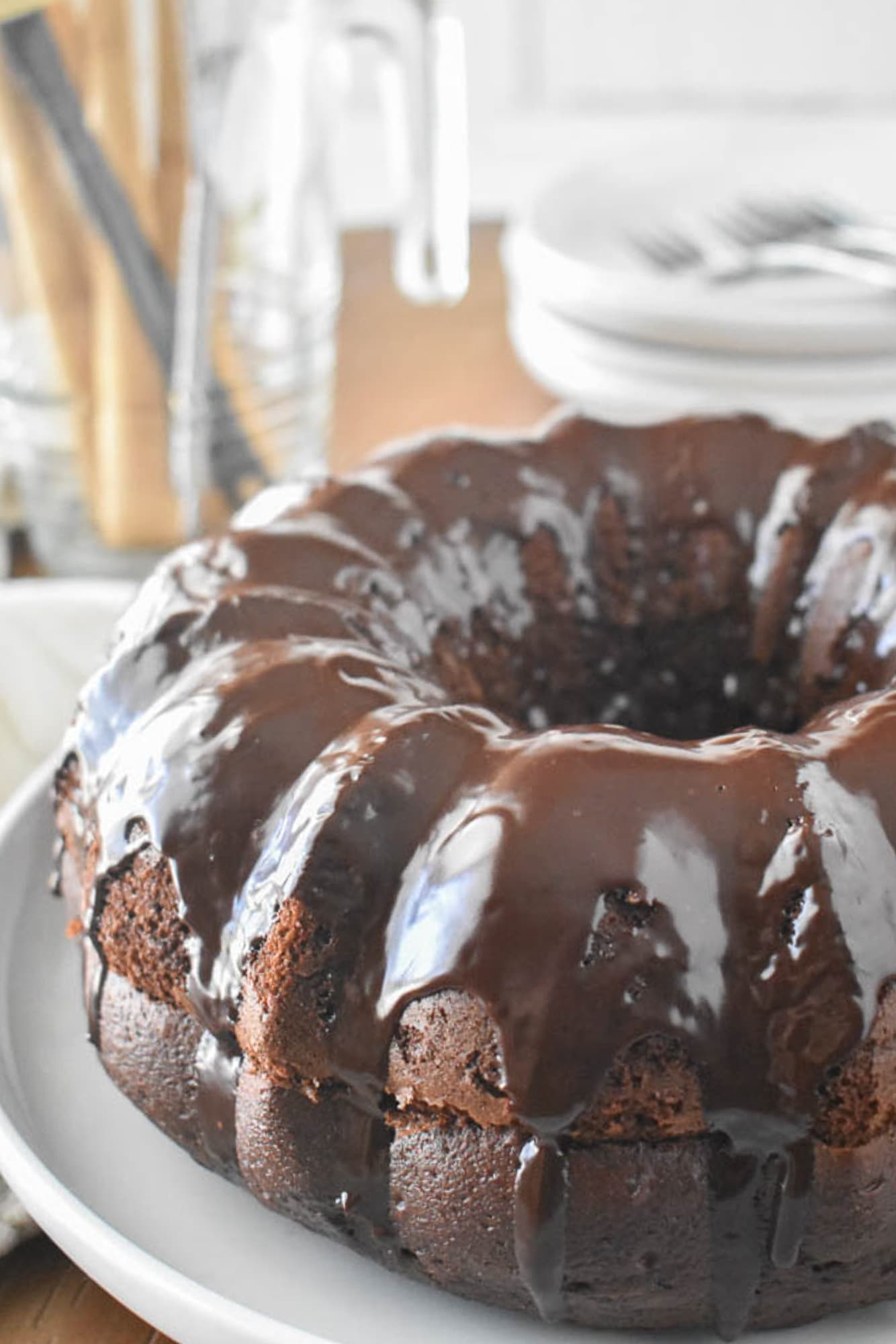 A chocolate Bundt cake (Black Russian Cake) with a glossy chocolate glaze sits on a white plate. The background is blurred, showcasing dishes and utensils.