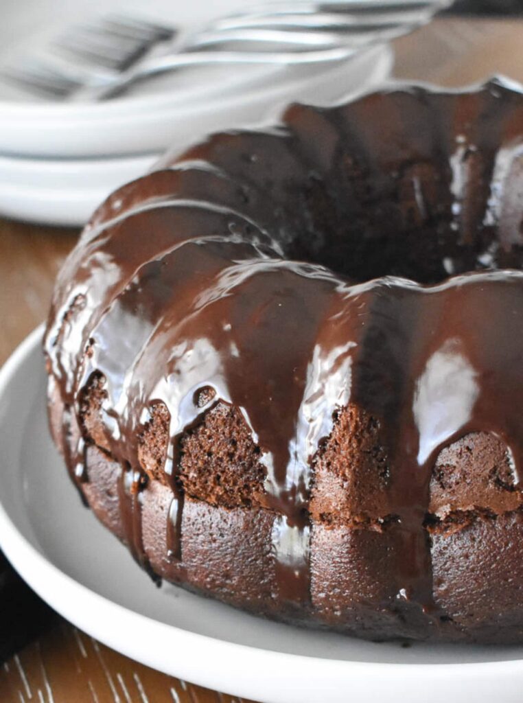 Rich chocolate Black Russian bundt cake with glossy chocolate glaze, presented on a white plate. The setting includes stacked plates and forks in the background
