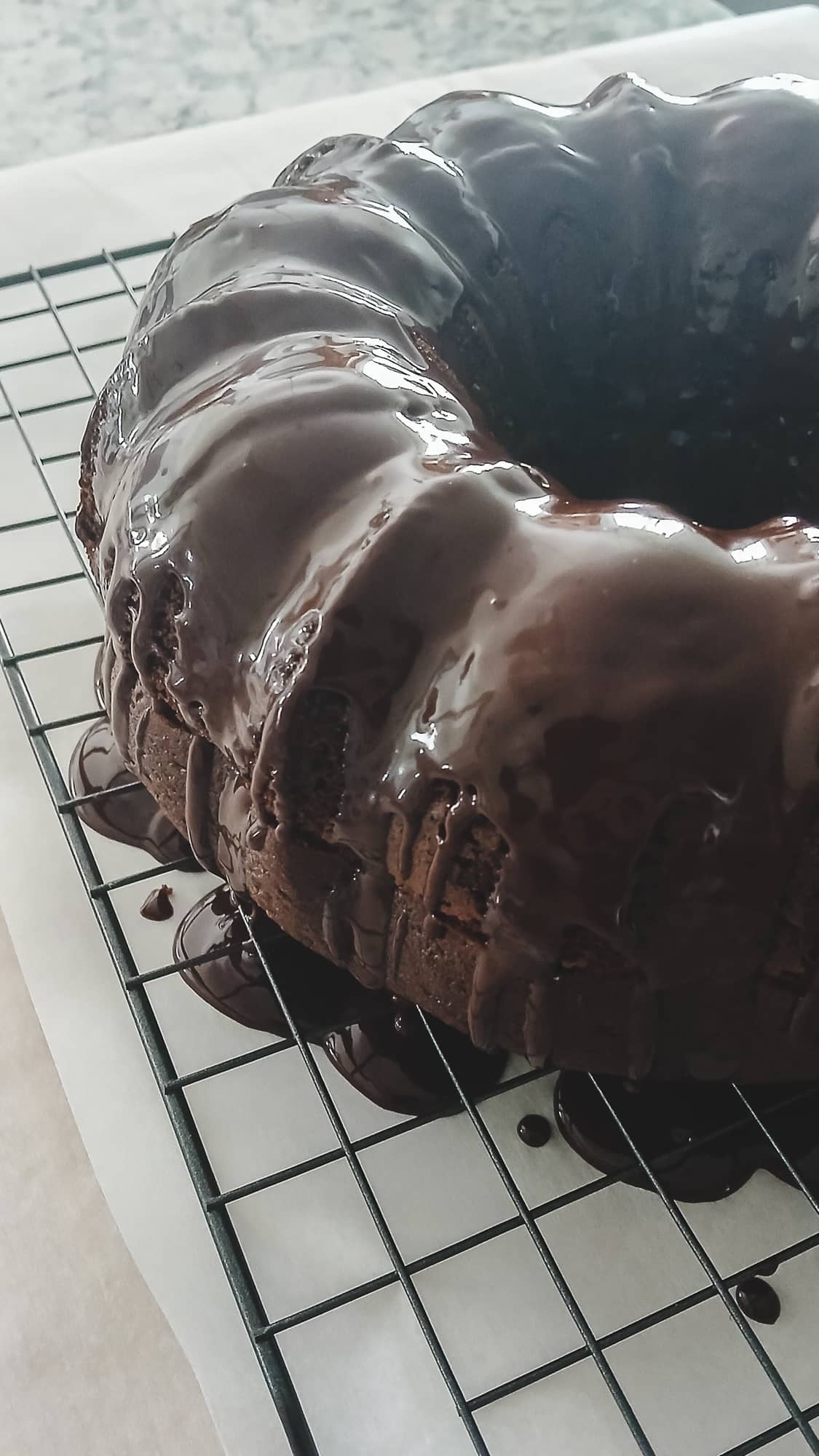 A rich chocolate Bundt cake on a cooling rack being covered in a glossy chocolate and espresso ganache glaze. The dark chocolate drips down the sides over parchment paper