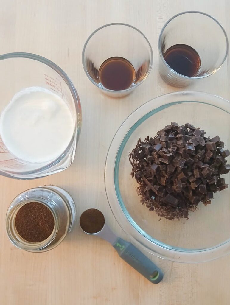 Chocolate ganache ingredients for Black Russian Cake: chopped chocolate in a bowl, a measuring cup of cream, two small glasses of liqueur, espresso powder jar, and teaspoon on wood surface.