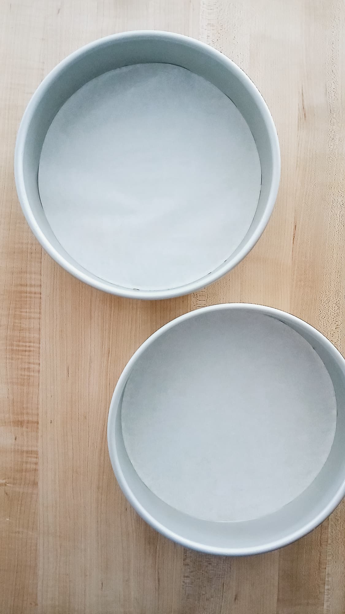 Two round cake pans lined with parchment paper sit on a wooden surface. The pans are empty and ready to fill with carrot cake batter