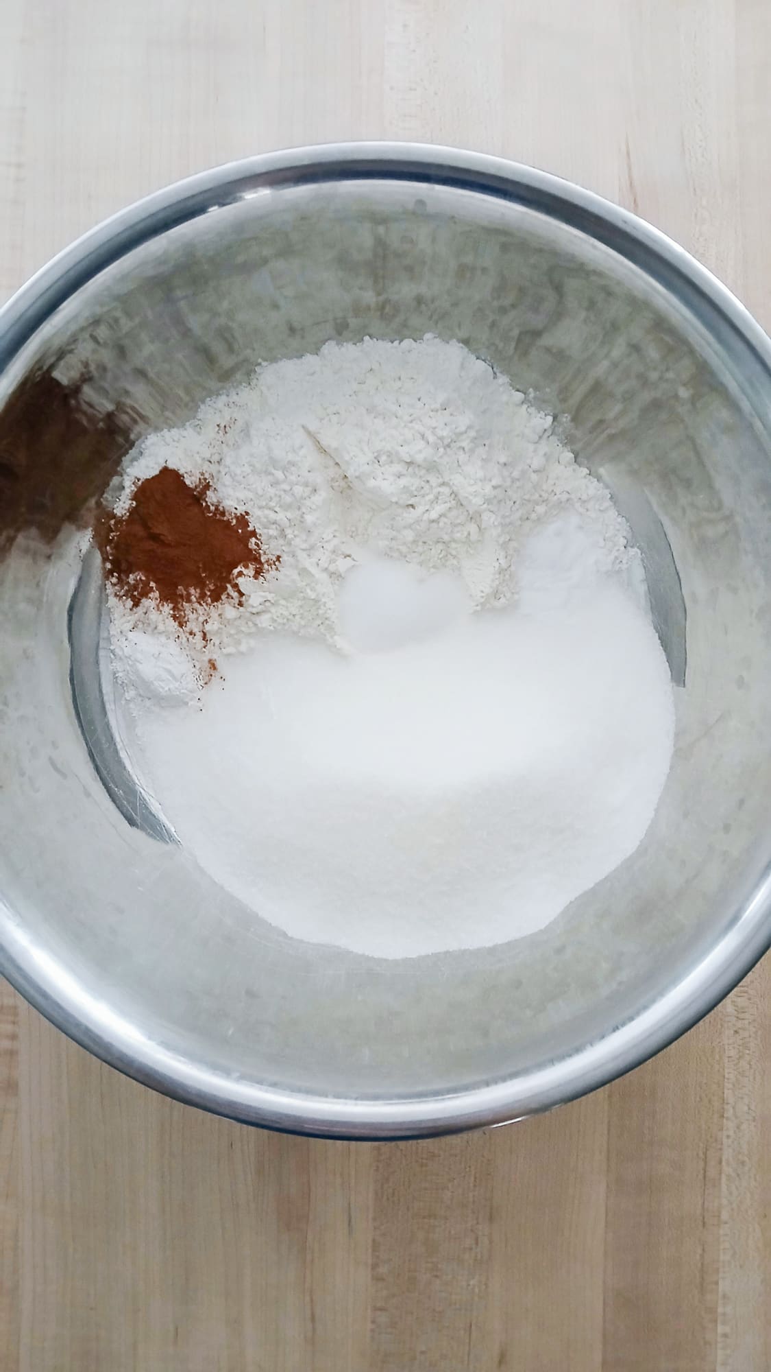 A metal bowl on a wooden surface contains sugar, flour, baking soda, baking powder, salt, and cinnamon ready for mixing.