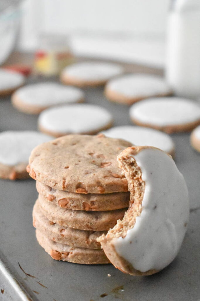 Stack of round cookies with a white frosting edge, one cookie partially bitten. Background shows more frosted cookies on a baking sheet. Cozy and inviting mood.