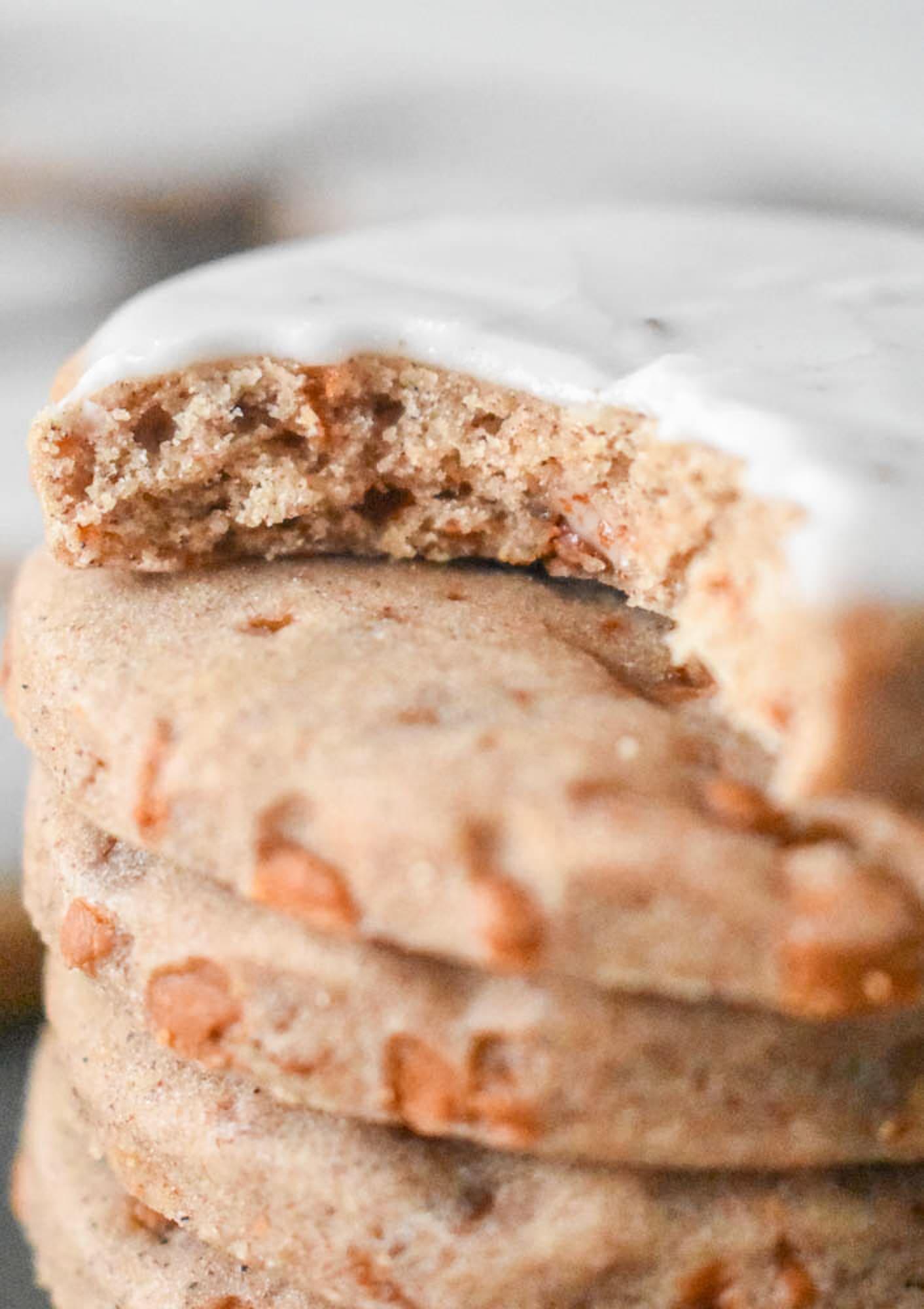 Close-up of a stack of chai sugar cookies topped with white glaze. The top cookie has a bite taken out, showing a soft interior and cinnamon chips on the inside.