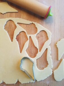 A cookie cutter shaped like an ice cream cone, resting on a sheet of sugar cookie dough with a rolling pin nearby
