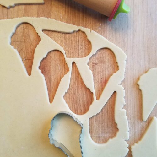 A cookie cutter shaped like an ice cream cone, resting on a sheet of sugar cookie dough with a rolling pin nearby