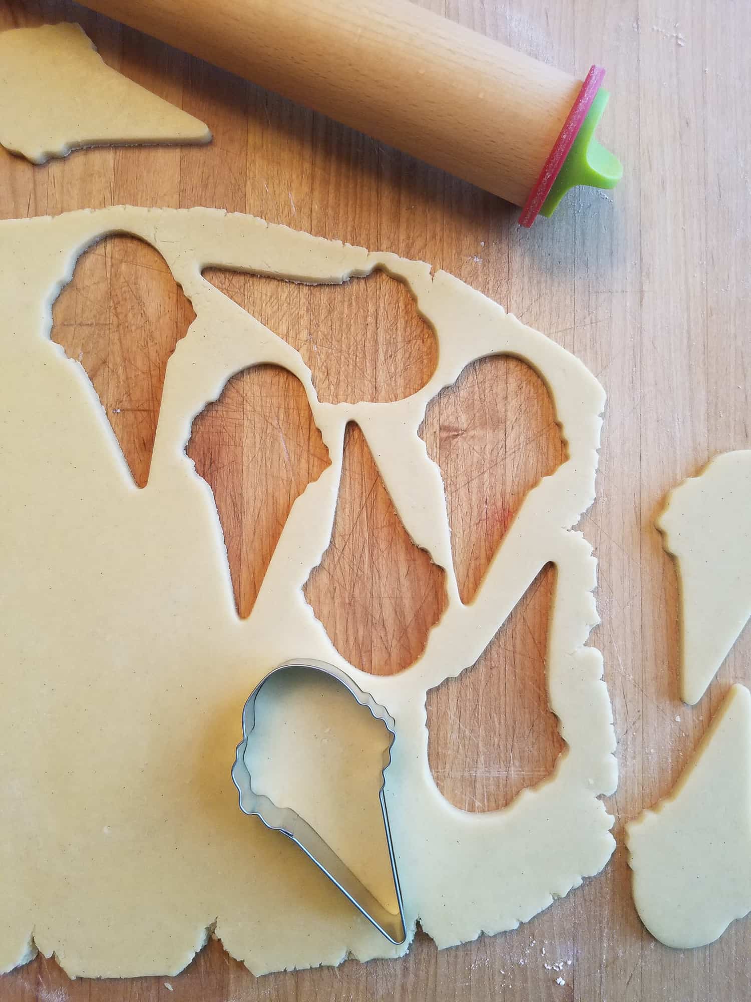 A cookie cutter shaped like an ice cream cone, resting on a sheet of sugar cookie dough with a rolling pin nearby