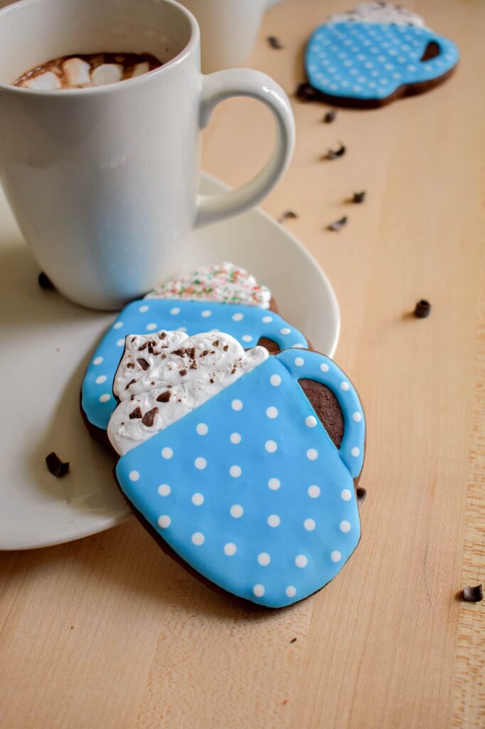 Blue polka dot mug-shaped cookies with whipped cream decoration made with royal icing. A mug of hot chocolate with marshmallows sits nearby
