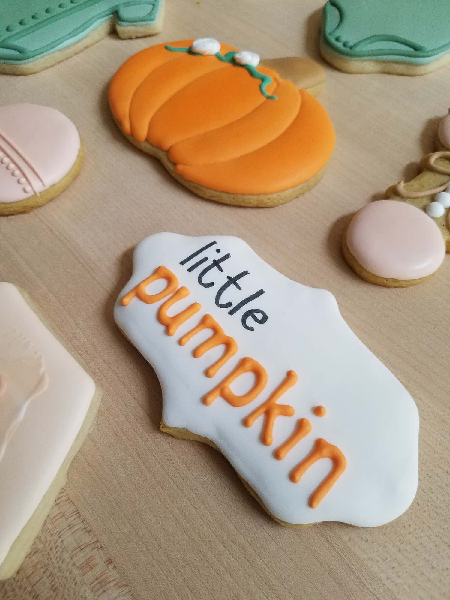 Cookies decorated using royal icing with meringue powder on a wooden surface. One cookie says "little pumpkin" in orange and black lettering, surrounded by autumn-themed shapes.