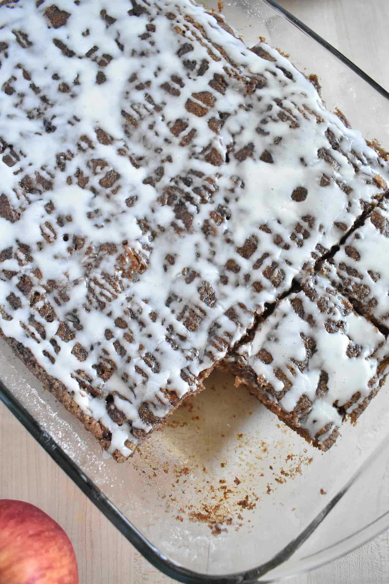 Spiced apple cake with a white icing drizzle pattern in a glass dish, partially sliced. A red apple is visible on the wooden table.