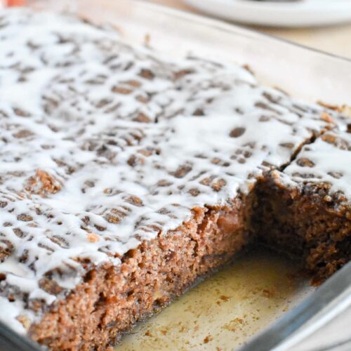 A glass dish holds a rectangular spiced apple cake drizzled with white glaze; a slice is missing. Two red apples and a plate with a piece are in the background.