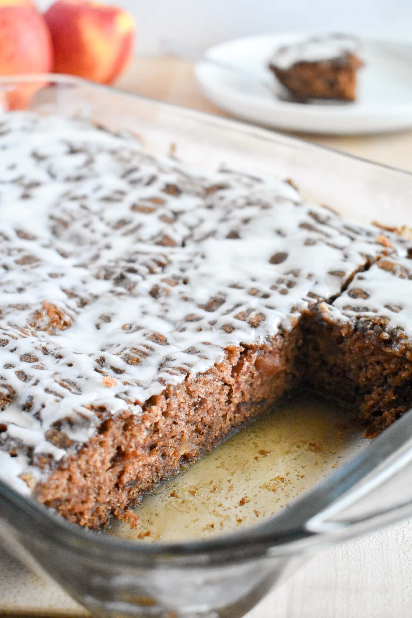 A glass dish holds a rectangular spiced apple cake drizzled with white glaze; a slice is missing. Two red apples and a plate with a piece are in the background.