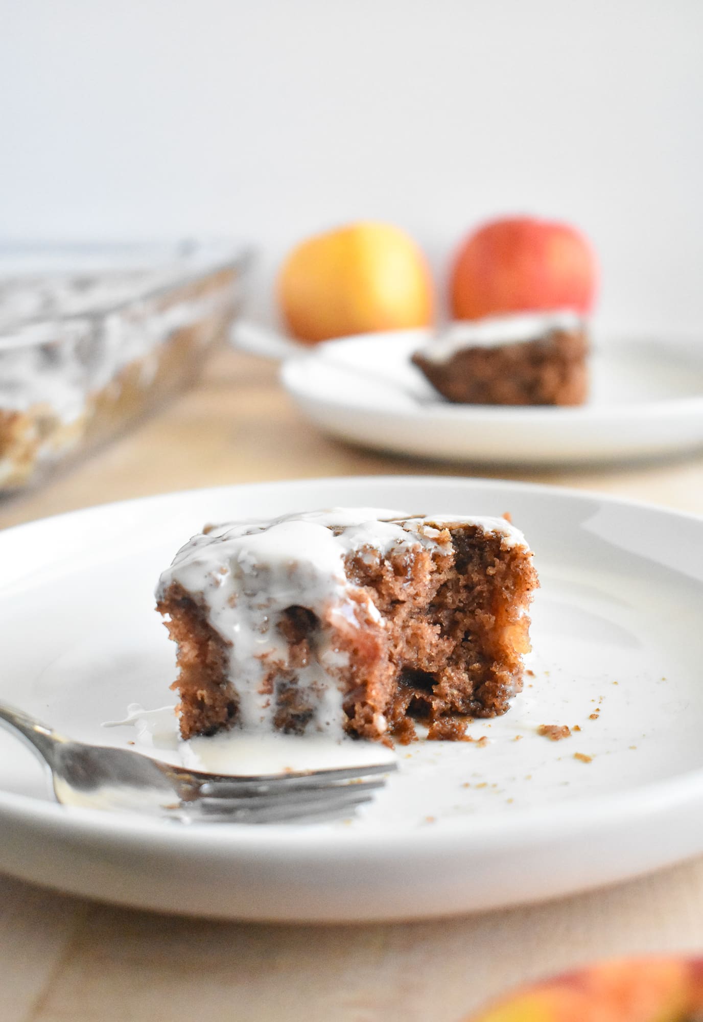 A close-up of a partially eaten slice of spiced apple cake with creamy icing on a white plate. Fork beside it, with blurred apples and another plate in the background.