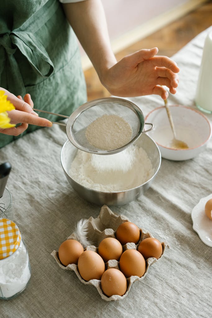 A person sifts flour surrounded by eggs, flour, and baking tools, creating a cozy baking scene.